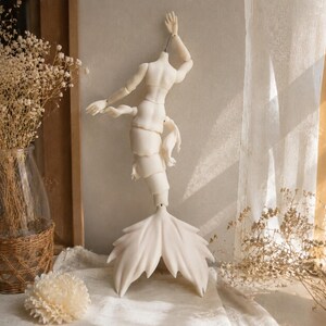 May include: A pale, articulated ball-jointed doll featuring a human-like upper torso with arms and a segmented, tail-like lower body ending in a wide, pointed fin. The doll is suspended by a metal hook at the neck, displaying a smooth, matte finish.