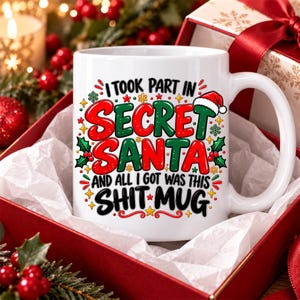Może przedstawiać: Biały ceramiczny kubek z napisem I TOOK PART IN SECRET SANTA AND ALL I GOT WAS THIS SHIT MUG w czerwonych i czarnych literach. Wzór zawiera zielone liście ostrokrzewu, czerwone jagody, małe żółte gwiazdki oraz czerwoną czapkę Świętego Mikołaja.