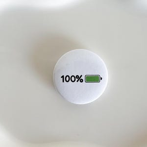 May include: A circular white pinback button featuring the black text 100% followed by a graphic of a battery icon with a green fill, indicating a full charge. The design is centered on the smooth, matte surface of the round button accessory.