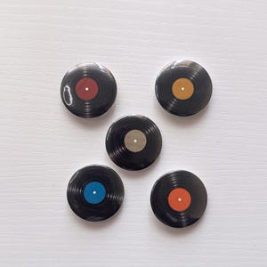May include: Five circular pinback buttons designed to resemble miniature vinyl records. Each button features a black grooved surface with a central label in a different solid color: red, gold, silver, blue, and orange, each with a small white center dot.