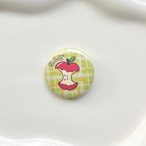 May include: A circular pinback button featuring a light green and white plaid background with a central illustration of a red apple core. The design includes small orange stars and the Japanese text "りんご" written in a rounded, white font.