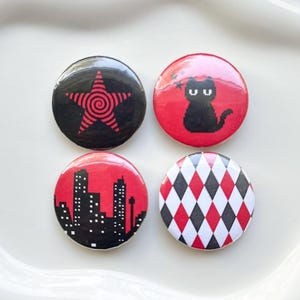 May include: Four circular pin-back buttons. The first is black with a red spiral star. The second is red with a black pixelated cat. The third is red with a black city skyline silhouette. The fourth features a red, black, and white diamond pattern.