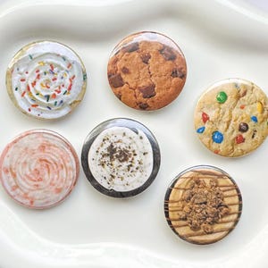 May include: A set of six circular pin-back buttons featuring realistic food imagery. The designs include a cupcake with sprinkles, a chocolate chip cookie, a cookie with colorful candy pieces, a pink swirl, a white cream top, and a peanut butter treat.