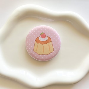 May include: A circular pinback button featuring a light pink background with white polka dots. The center displays a tan-colored custard dessert topped with a dollop of white whipped cream and a single small red strawberry with green leaves.