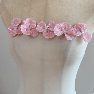 May include: A strapless garment featuring a horizontal row of six dimensional, light pink floral appliqués. Each flower is crafted with dense, shimmering sequins and small white beads at the center, creating a textured, reflective surface across the chest.