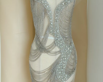 May include: A silver beaded embellishment with a chain-like design. The embellishment is on a white dress form and appears to be made of rhinestones.