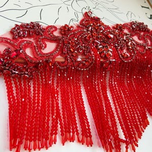Può includere: Primo piano di un bordo di perline rosse con ricami floreali intricati. Il bordo presenta una delicata base a rete, impreziosita da perline e paillettes rosse. Lunghe ciocche di perline rosse pendono dal basso, creando un effetto frangia. Questo bordo è adatto per abbellire capi d'abbigliamento.