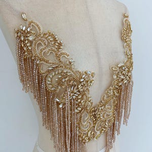 Puede incluir: Aplique bordado con cuentas doradas y cristales con flecos. El aplique presenta intrincados diseños florales y de volutas, adornados con cristales transparentes y flecos de cuentas colgantes. Se muestra en un maniquí, ideal para diseño de prendas.