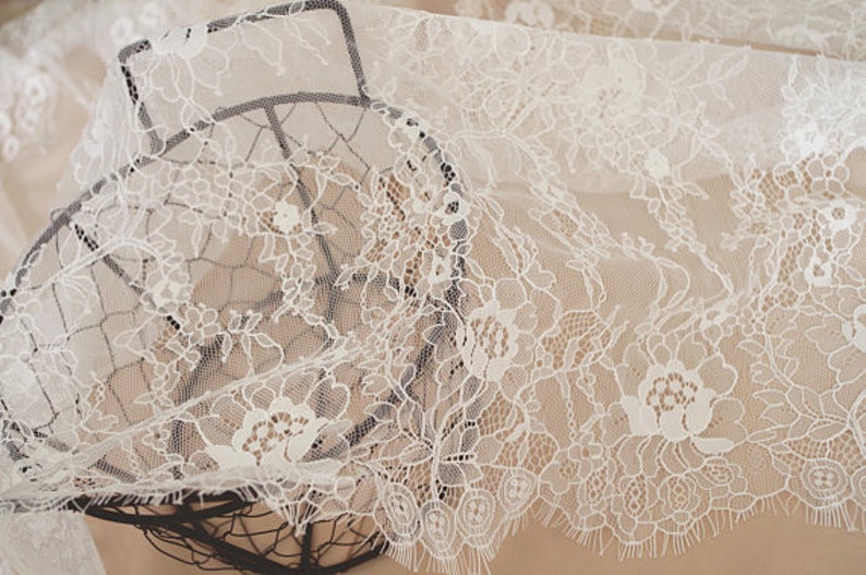May include: White lace fabric with a delicate floral pattern. The lace is draped over a wire basket.