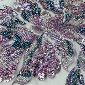 May include: A sheer tulle fabric appliqué featuring intricate floral embroidery embellished with iridescent lavender, silver, and deep teal glass beads and sequins. The design showcases organic petal shapes with dense beadwork and metallic thread detailing.