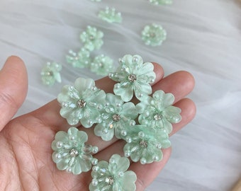 May include: A collection of light green fabric flowers with pearl and rhinestone embellishments. The flowers are approximately 1 inch in diameter.