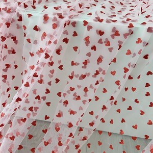 May include: Pink tulle fabric with red glitter hearts printed on it. The fabric is folded and draped over a white surface.