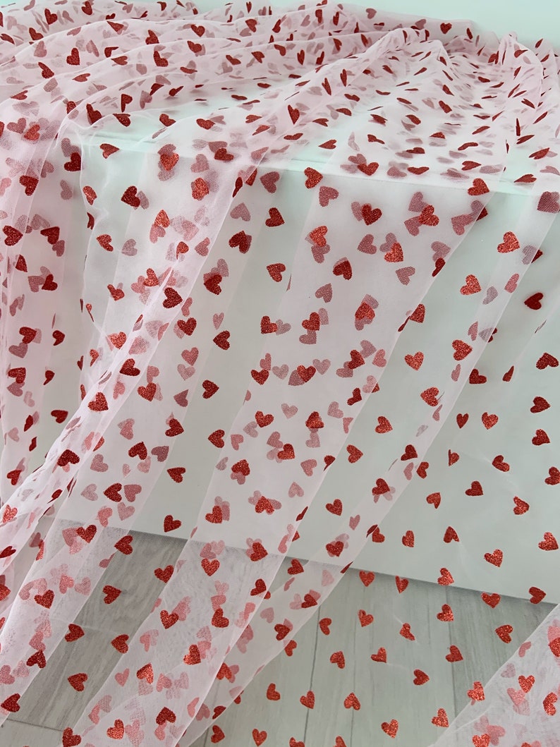 May include: Pink tulle fabric with red glitter hearts printed on it. The fabric is folded and draped over a white surface.
