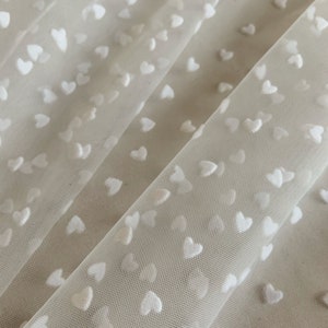 Pode incluir: Tecido de tule branco com um padrão de pequenos corações brancos. O tecido é transparente e delicado, e os corações são em relevo e texturizados.