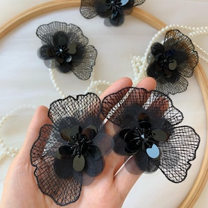 May include: Close-up of several black floral embellishments. Each flower features a layered design with a mesh overlay, sequins, and beaded centers. Some are arranged on a wooden hoop with a pearl strand.