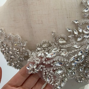 Puede incluir: Primer plano de una tela blanca con un ribete decorativo de pedrería y cuentas transparentes. El ribete tiene un patrón floral y está cosido a la tela.