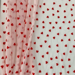 Op de afbeelding: Roze en witte doorschijnende stof met rode hartvormige patronen. De stof is gelaagd en de harten zijn aan beide kanten zichtbaar.