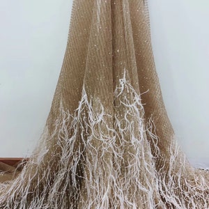 Puede incluir: Tela beige con ribete de plumas blancas. La tela tiene una textura brillante y reluciente.