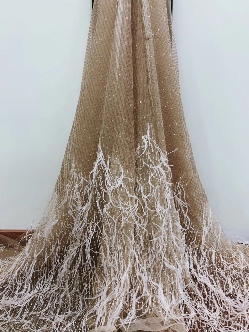 Puede incluir: Tela beige con ribete de plumas blancas. La tela tiene una textura brillante y reluciente.