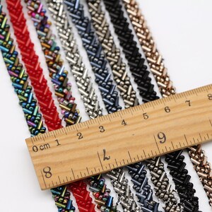 May include: Assortment of beaded trim in various colors, including red, black, silver, and iridescent. The trim is arranged in rows on a wooden ruler marked in centimeters and inches.