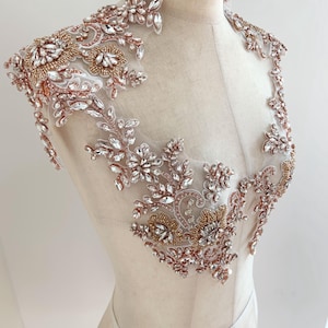May include: A rose gold and clear crystal beaded applique, with floral and leaf patterns. The applique is attached to a sheer, light-coloured fabric, creating a delicate embellishment. Suitable for bridal wear or evening gowns.