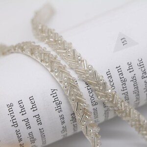 May include: A close-up of two strands of silver beaded trim. The trim is made of small, round beads and long, thin beads arranged in a chevron pattern.