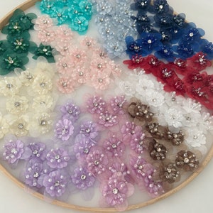 Puede incluir: Surtido de flores de tela de colores con detalles de perlas y pedrería. Las flores están dispuestas en círculo sobre un fondo blanco.