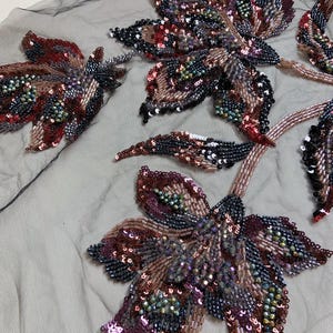 Puede incluir: Una tela de tul negro transparente con intrincados bordados florales adornados con lentejuelas y cuentas de vidrio en tonos rojo oscuro, cobre y negro. El diseño muestra pétalos y tallos detallados dispuestos en un patrón botánico.