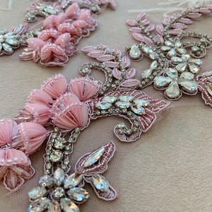 Puede incluir: Dos aplicaciones florales decorativas con lentejuelas rosa claro, cuentas y cristales facetados transparentes dispuestos en patrones de hojas y pétalos sobre una base de malla transparente. El bordado intrincado incluye hilos metálicos plateados.