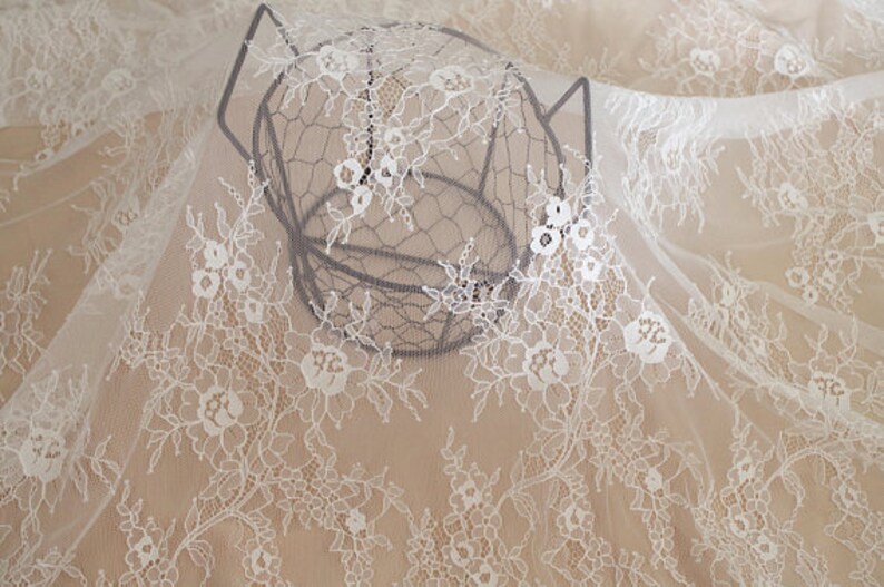 May include: White lace fabric with a delicate floral pattern. The fabric is draped over a wire basket.
