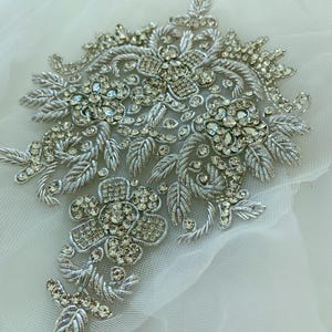 以下が含まれることがあります： 透明なクリスタルラインストーンが付いた、銀色の刺繍入りフローラルアップリケ。アップリケは、衣服やアクセサリーを飾るのに最適な、詳細な葉と花の模様が特徴です。複雑なビーズ細工がエレガントさと輝きを添えています。
