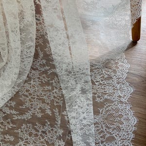 May include: Close-up of delicate white lace fabric. The lace features intricate floral and leaf patterns, with a scalloped edge. The fabric is semi-sheer, allowing light to pass through, and is likely used for bridal wear or home decor.