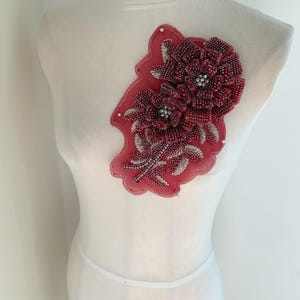 May include: A decorative applique featuring two beaded flowers in shades of burgundy and silver. The flowers are attached to a red fabric backing with a scalloped edge. The applique is displayed on a white mannequin.