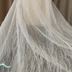 Puede incluir: Tela de tul blanco con una textura delicada y brillante y un dise&ntilde;o de flecos. La tela es transparente y tiene un brillo sutil.