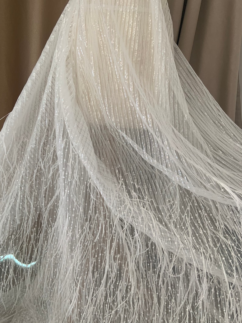 Puede incluir: Tela de tul blanco con una textura delicada y brillante y un dise&ntilde;o de flecos. La tela es transparente y tiene un brillo sutil.