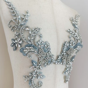 May include: A delicate, floral-inspired embellishment featuring intricate designs and sparkling rhinestones. The embellishment is made of a light blue fabric with silver accents and is perfect for adding a touch of elegance to any garment.