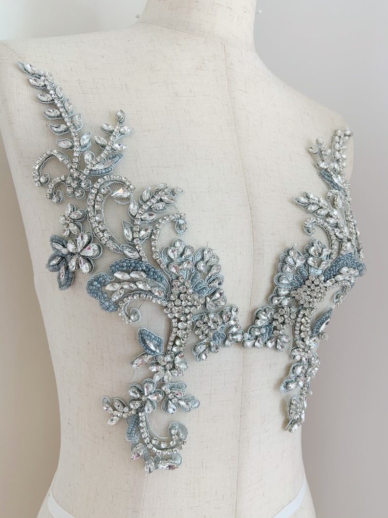 May include: A delicate, floral-inspired embellishment featuring intricate designs and sparkling rhinestones. The embellishment is made of a light blue fabric with silver accents and is perfect for adding a touch of elegance to any garment.
