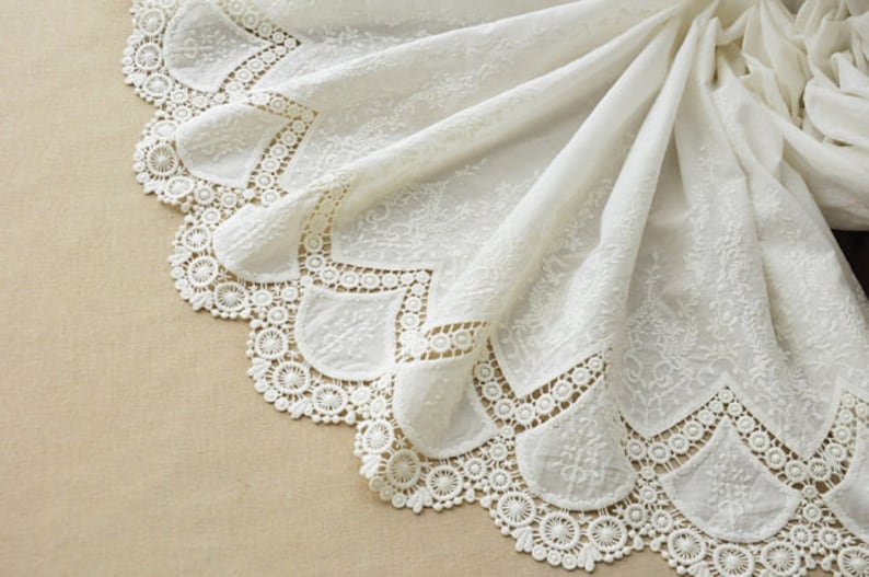 May include: White cotton fabric with a delicate lace trim. The lace features a scalloped edge with intricate floral designs.
