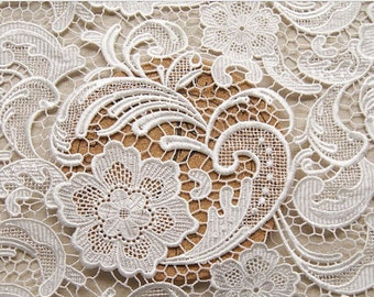 May include: White lace fabric with a delicate floral pattern. The lace features intricate swirls and a large flower in the center.