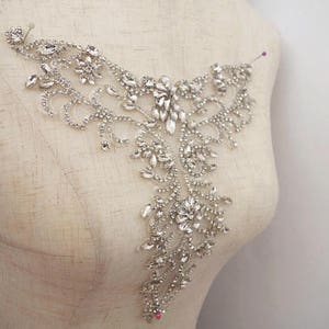May include: A silver rhinestone embellishment with a floral design. The embellishment is on a white fabric background.