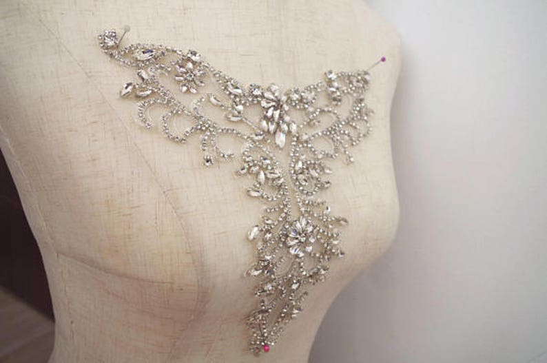 May include: A silver rhinestone embellishment with a floral design. The embellishment is on a white fabric background.
