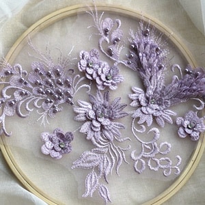 May include: A close-up of a purple floral lace applique with pearl and rhinestone embellishments. The applique is displayed on a wooden embroidery hoop.