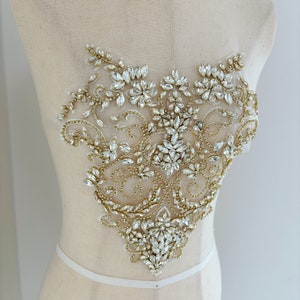 May include: A gold and clear crystal beaded applique with a floral design. The applique is on a white background.