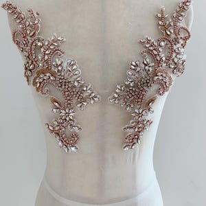 May include: A pair of symmetrical floral-shaped appliques featuring clear faceted crystals and rose gold metallic beads arranged on a sheer white mesh base. The intricate design includes leaf and vine motifs with dense beadwork and crystal accents.