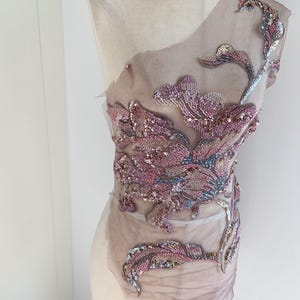 Puede incluir: Una tela transparente de color malva con intrincados bordados florales. El bordado presenta lentejuelas y cuentas rosas, moradas y plateadas, creando un diseño decorativo texturizado. La tela está drapeada sobre un maniquí, mostrando el potencial para el embellecimiento de prendas.