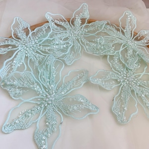 May include: Several light blue floral appliques with intricate bead and sequin detailing. The appliques are arranged on a wooden hoop, showcasing their delicate design. These embellishments are ideal for crafting and adding a touch of elegance.