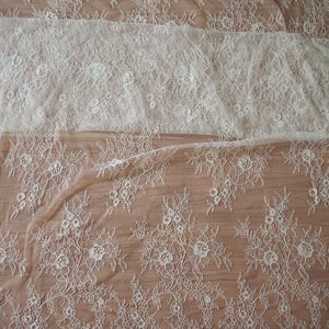 May include: White floral lace fabric with a delicate pattern. The lace has a scalloped edge and is sheer.