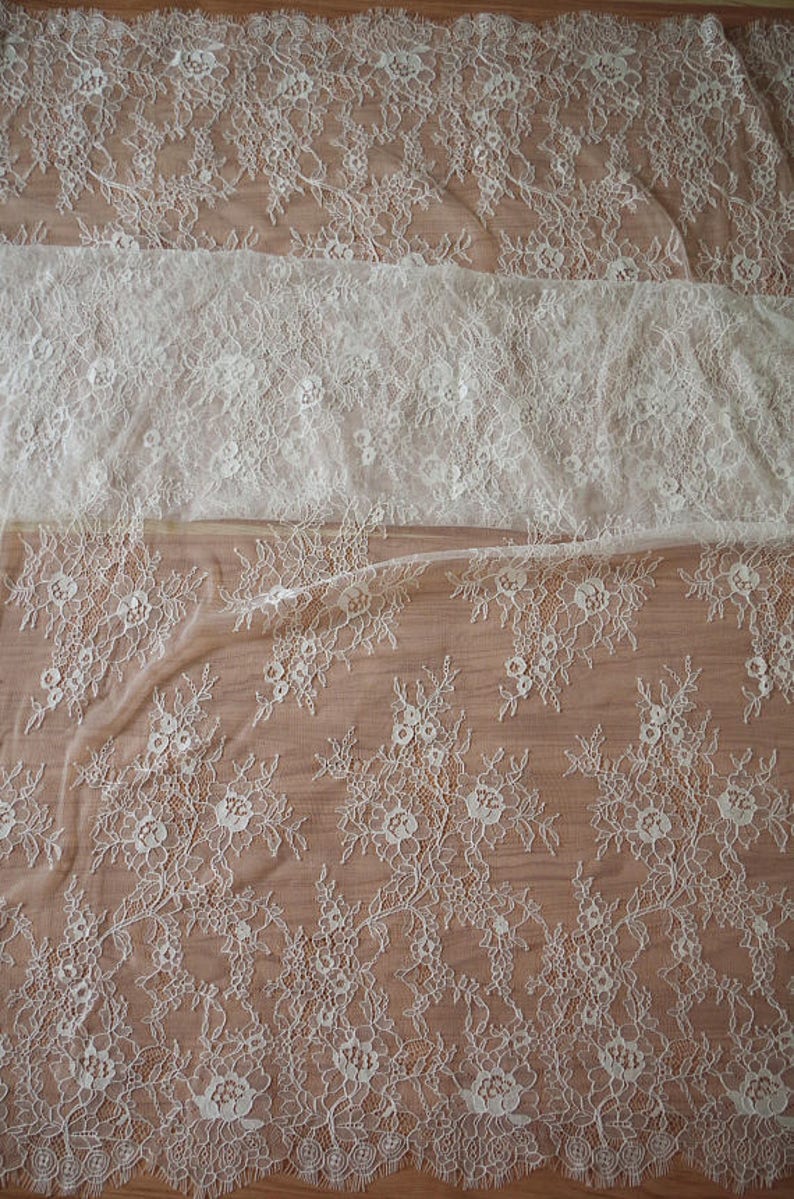 May include: White floral lace fabric with a delicate pattern. The lace has a scalloped edge and is sheer.