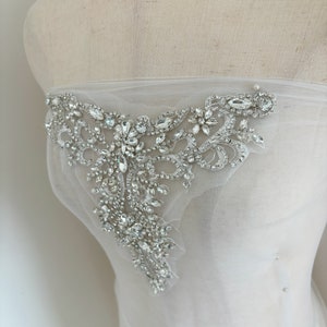 May include: A white tulle fabric with a large, ornate, crystal embellishment. The embellishment is made of silver and clear crystals and has a floral design.