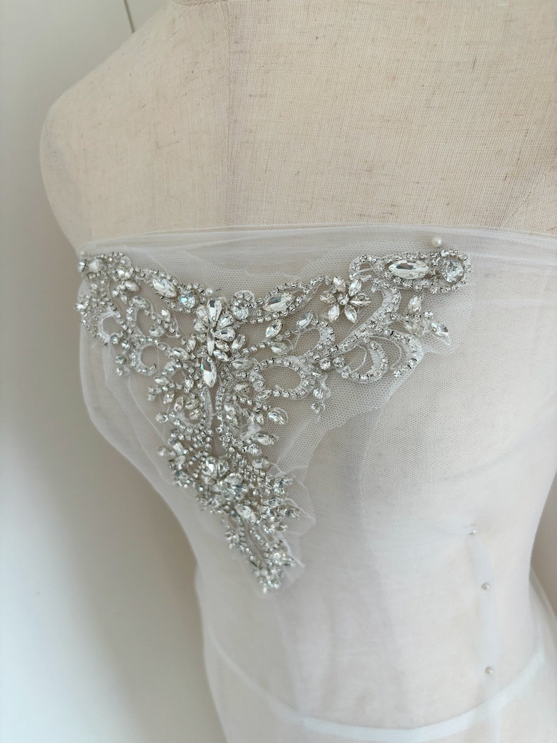 May include: A white tulle fabric with a large, ornate, crystal embellishment. The embellishment is made of silver and clear crystals and has a floral design.